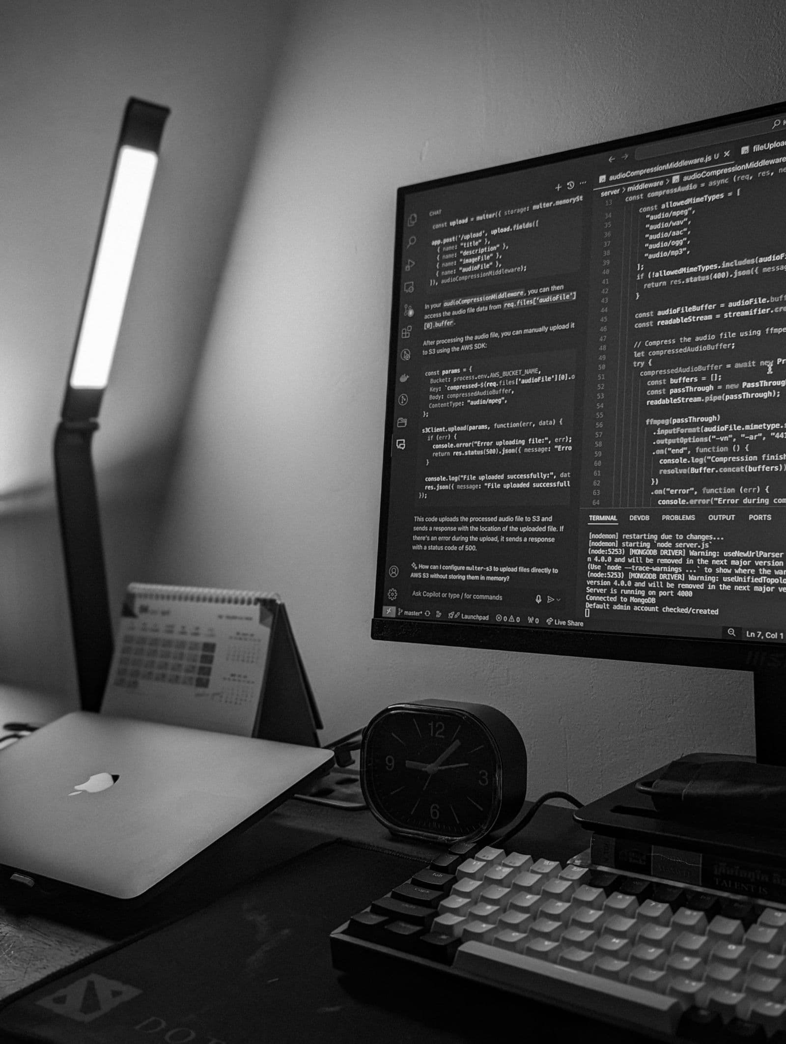 A monitor showing code beside a laptop and mechanical keyboard on a dark desk
